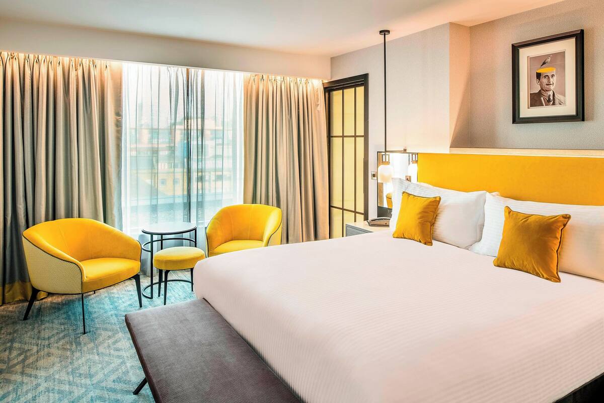 A hotel room with a large bed, two yellow armchairs, a small table, and a view of the city through large windows.