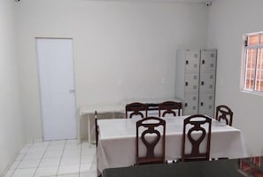 A dining area with a table set for four, surrounded by chairs with heart-shaped backrests. There are lockers against the wall and a door to the left.