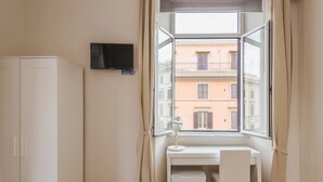 A room with a white desk, a chair, a TV mounted on the wall, and a window with a view of buildings. - Walker Rooms (Rome)