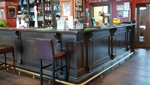 A bar area with a dark wood bar, two stools, and a variety of bottles and glasses on shelves. - Horse and Jockey Inn (Matamata)