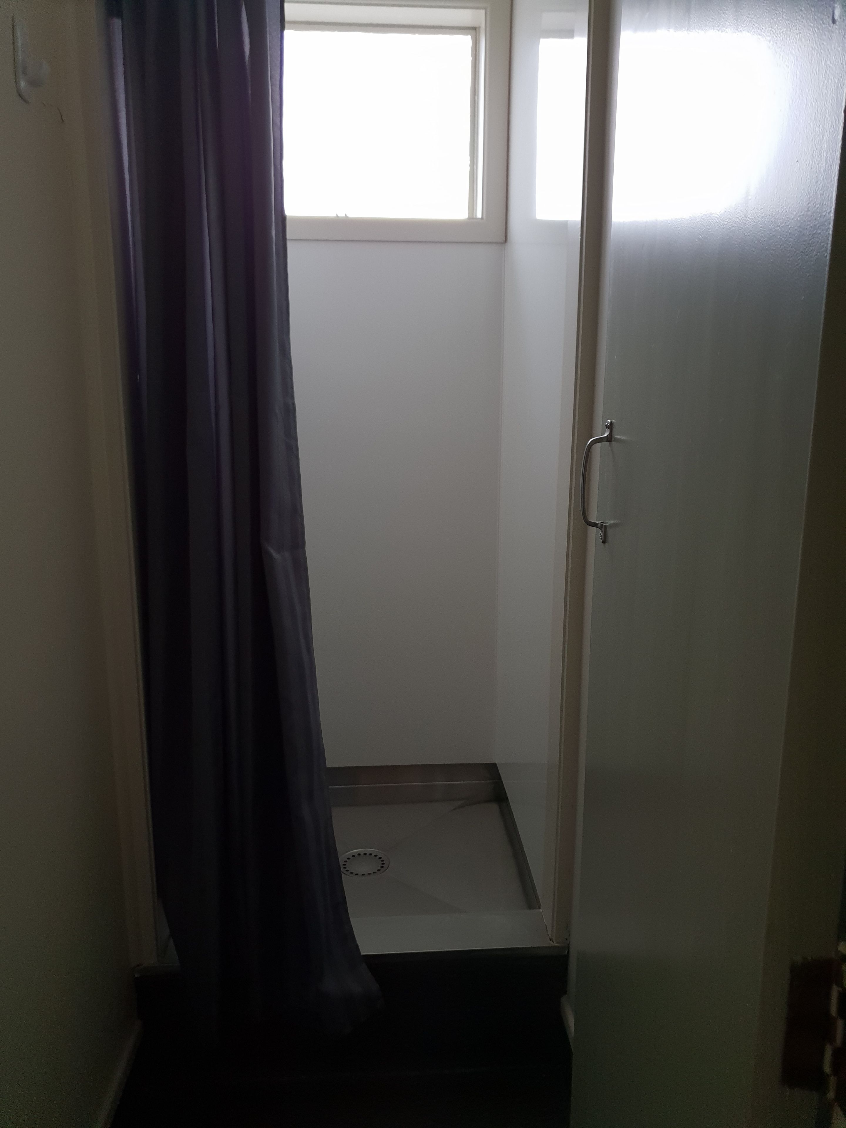 A small, narrow bathroom with a shower area, a door with a handle, and a window allowing natural light.