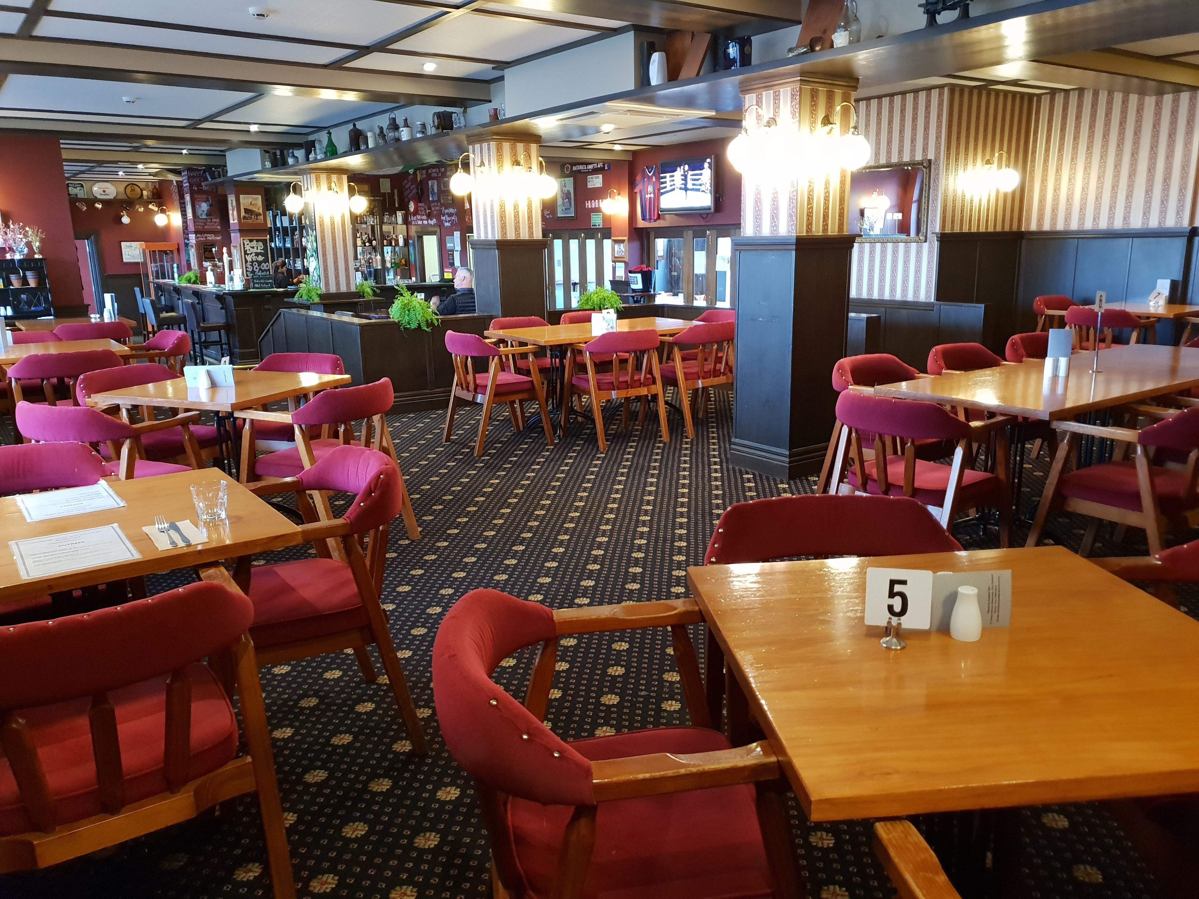 Un restaurante con mesas de madera y sillas rojas, alfombra estampada y una zona de bar al fondo.