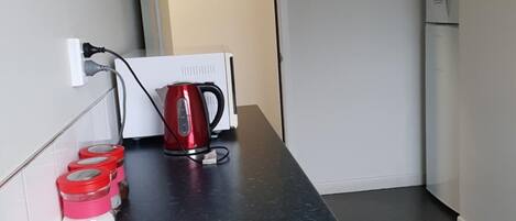 Cozinha pequena com bancada, chaleira e máquina de café. Há uma pia com escorredor e uma porta que dá para outro cômodo.