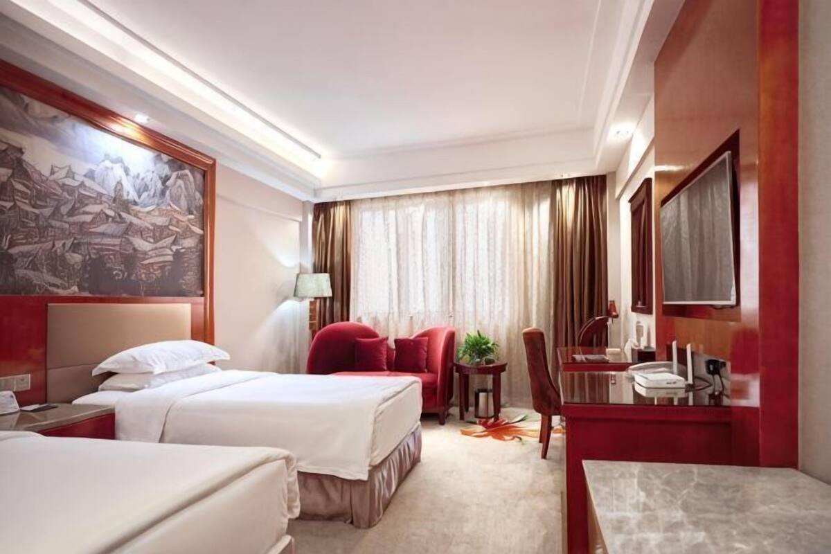 A hotel room with two beds, a desk, a chair, a small table, a mirror, and a window with curtains.