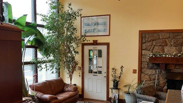 A spacious living room with a brown leather sofa, a green patterned rug, a stone fireplace, and a large window with a view of a building.