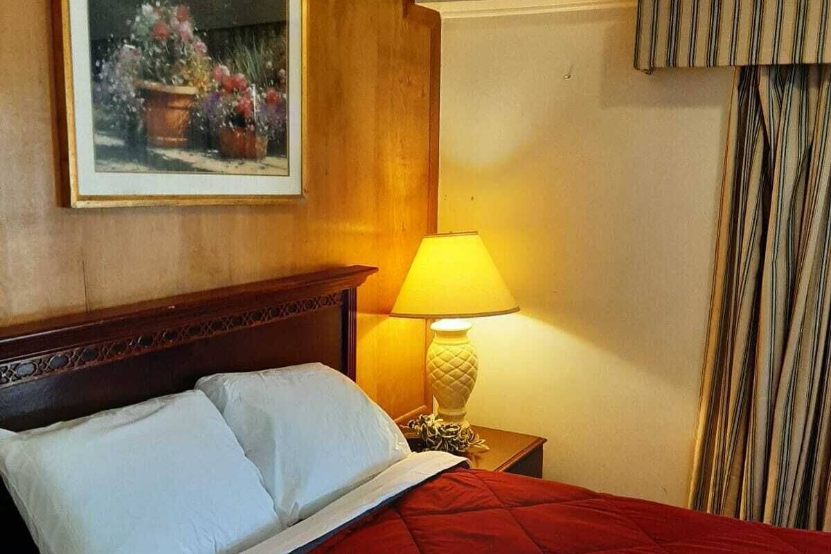 Una habitación de hotel con cabecera de madera, cama con ropa de cama blanca y roja, un cuadro de flores en la pared, una lámpara en una mesita de noche y cortinas a rayas.