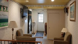 A hotel lobby with a reception desk, a waiting area with chairs, a door leading to the hallway, and a sign with a phone number.