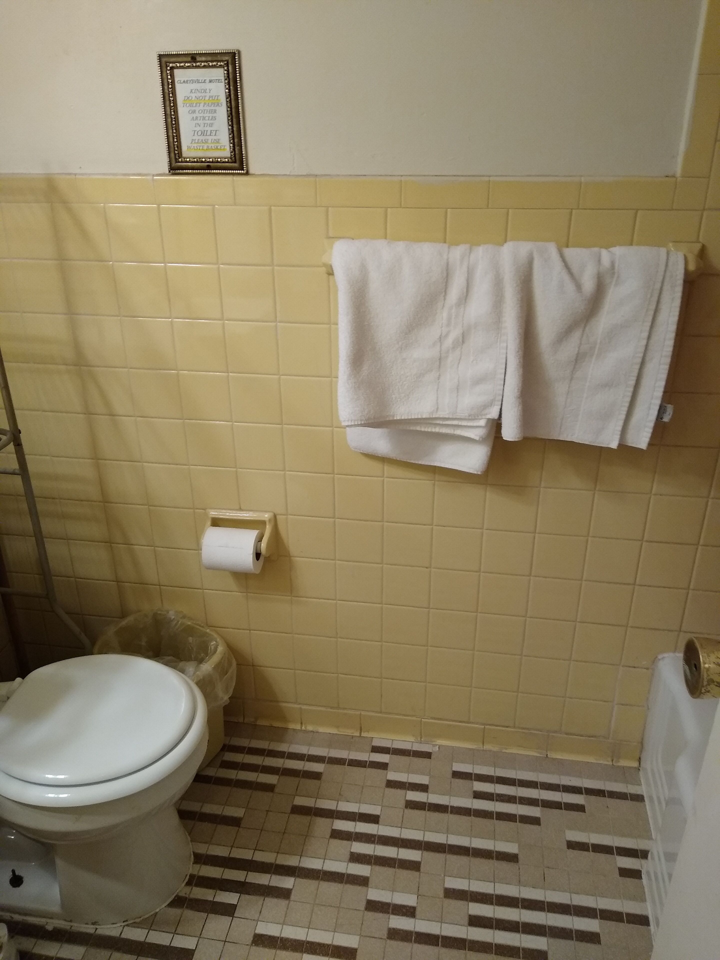 Une salle de bain équipée de toilettes, d’une poubelle, d’un porte-serviettes avec deux serviettes et d’un panneau encadré accroché au mur.