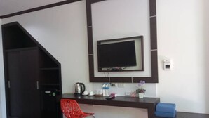 Una habitación con un escritorio marrón oscuro, un televisor de pantalla plana montado en la pared, una silla roja, un cojín azul, un hervidor de agua, una botella de agua y una pequeña planta en maceta.