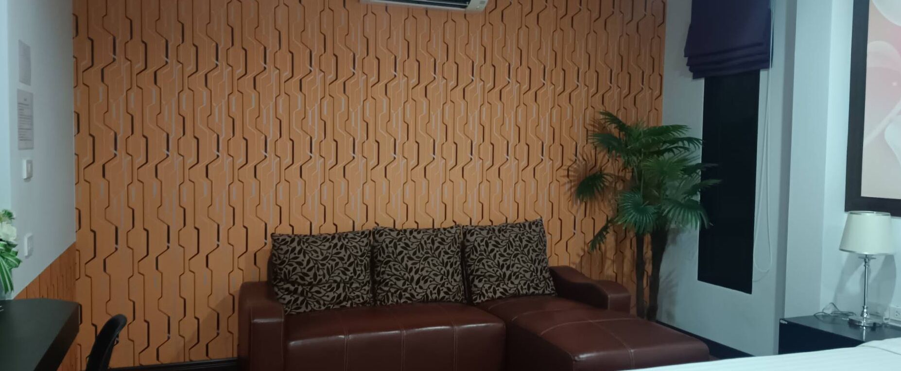Een kamer met een behang, een bruine leren bank met decoratieve kussens, een bijzettafeltje met een lamp en een potplant.