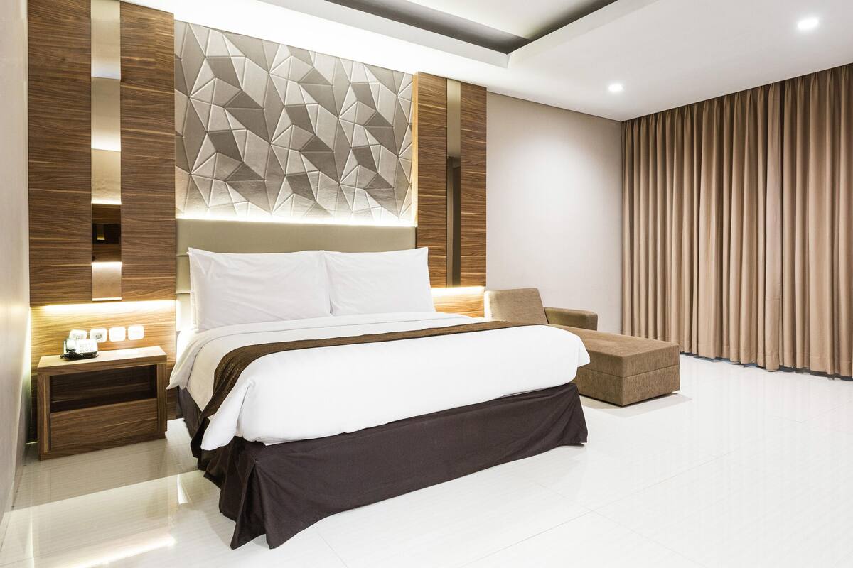 A modern hotel room with a large bed, a bedside table with a lamp, a sofa, and a large window with curtains.