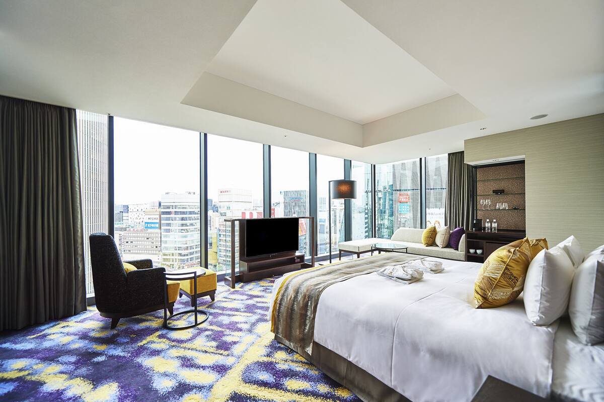 A hotel room with a large bed, a TV, a desk, and a view of the city.