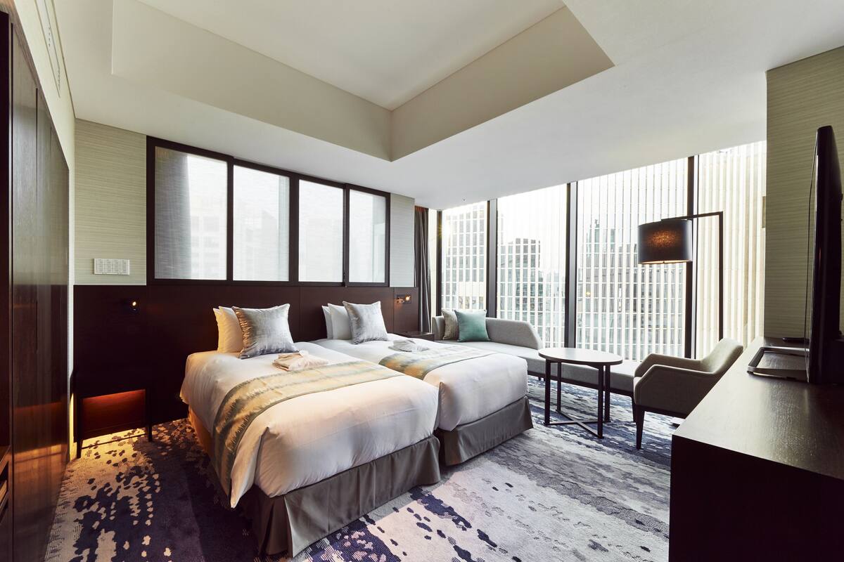 A hotel room with a large bed, a desk, and a view of the city through large windows.