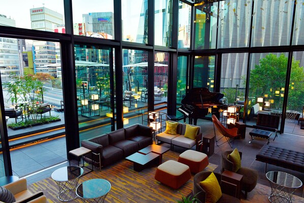 A modern lounge with a city view, featuring sofas, tables, and a piano.