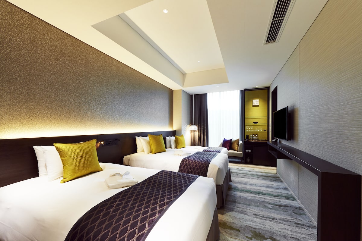 A hotel room with two beds, a desk, a flat-screen TV, and a minibar.