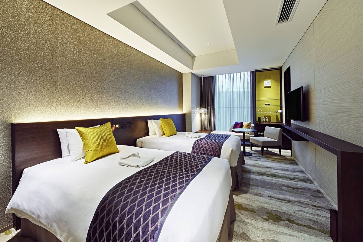 A hotel room with two beds, a desk, a chair, a TV, and a bathroom.