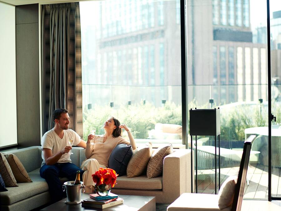 Una sala de estar moderna con un sofá, una mesa de centro con un jarrón de flores y vistas a un paisaje urbano a través de amplios ventanales.