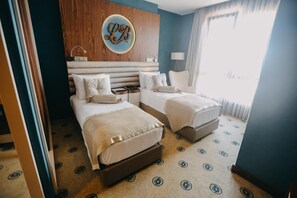 Habitación de hotel con dos camas, cabecero de madera, decoración circular en la pared, mesita de noche con lámpara y ventana con cortinas.