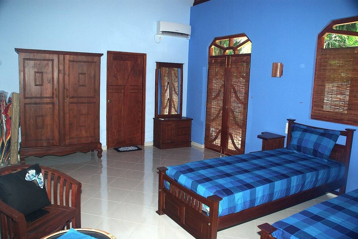 A bedroom with a bed, a wooden wardrobe, a small cabinet, and a window with a view of greenery.