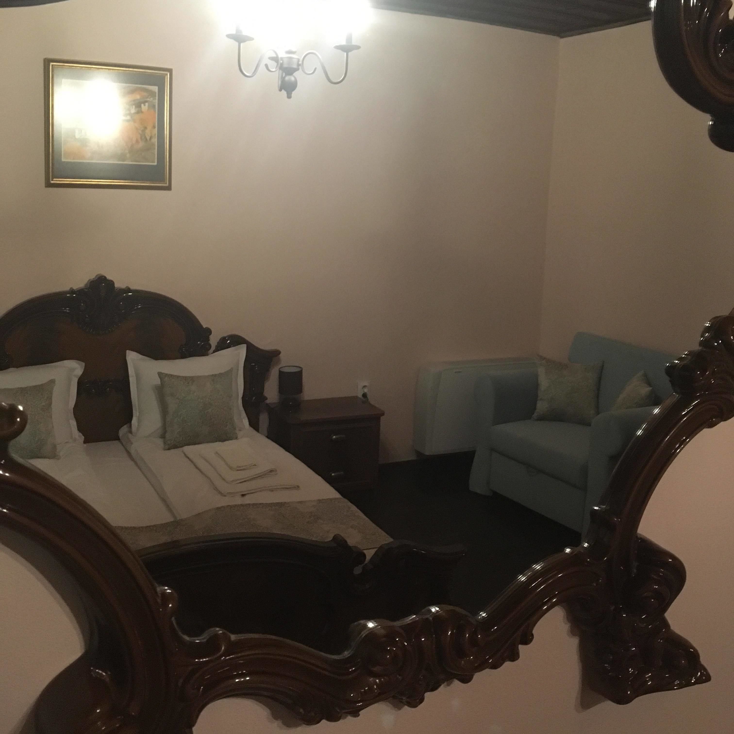 A bedroom with a large ornate mirror, a bed with pillows, a nightstand, a chair, and a wall-mounted light fixture.