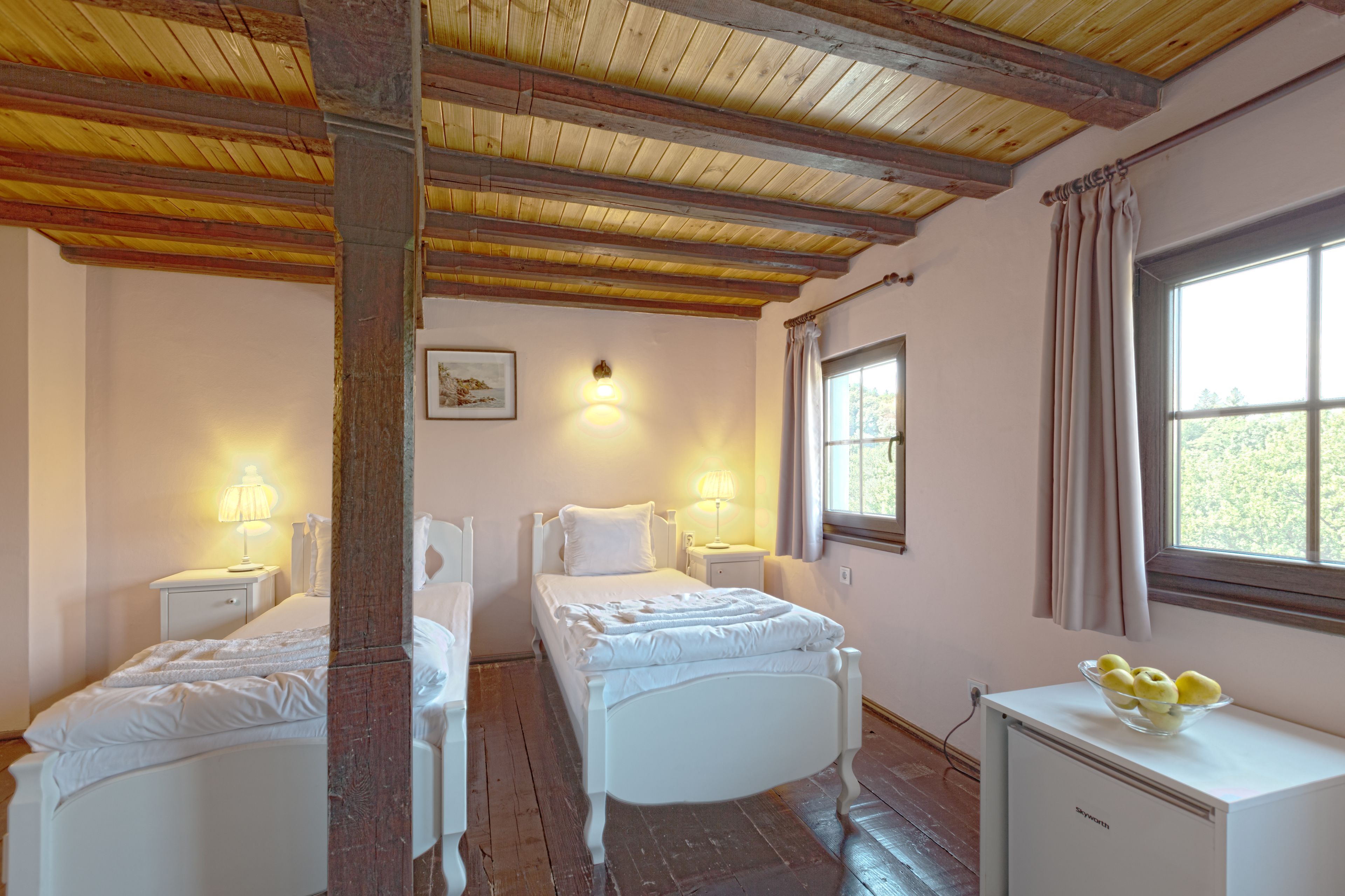 A bedroom with two beds, wooden beams on the ceiling, bedside lamps, a window with curtains, and a small cabinet with fruit.
