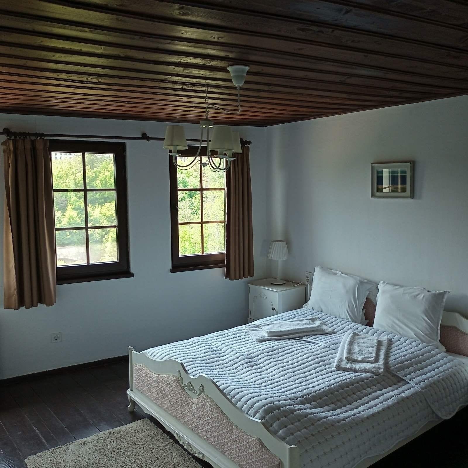 Un dormitorio con cama, dos ventanas, una mesita de noche y una lámpara.