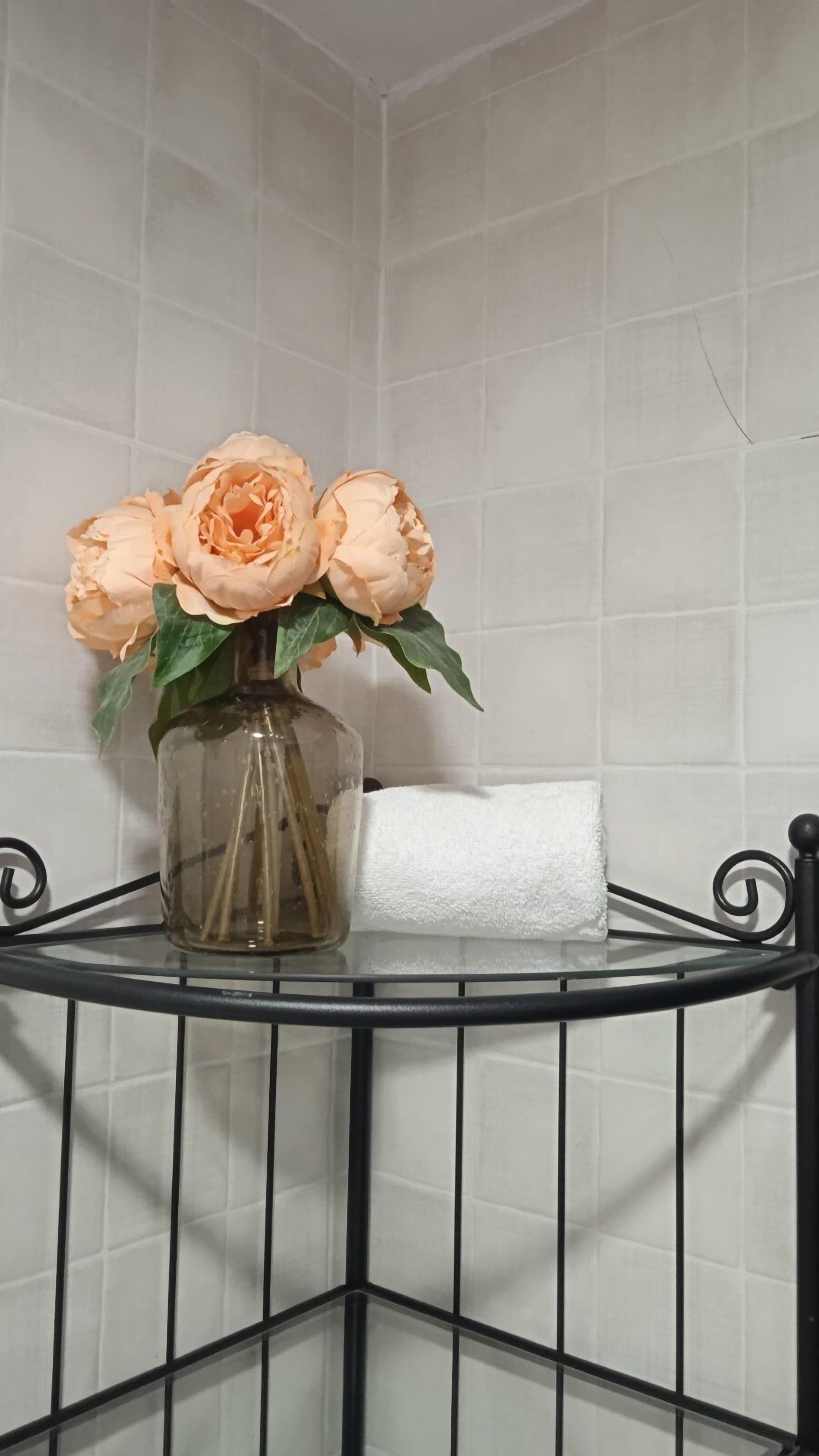 Un florero de vidrio con flores sobre una repisa metálica en un rincón de una habitación con paredes de azulejos blancos.