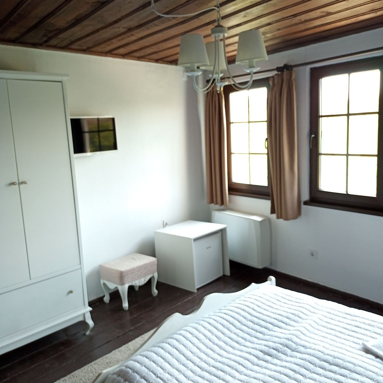 A bedroom with a bed, a wardrobe, a small table, a window with curtains, and a ceiling fan.
