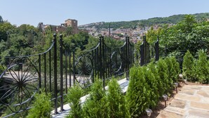 A stone pathway leading to a metal fence with decorative elements, surrounded by greenery and overlooking a cityscape with buildings and hills.