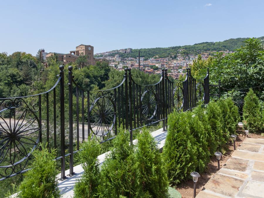 A stone pathway leading to a metal fence with decorative elements, surrounded by greenery and overlooking a cityscape with buildings and hills.