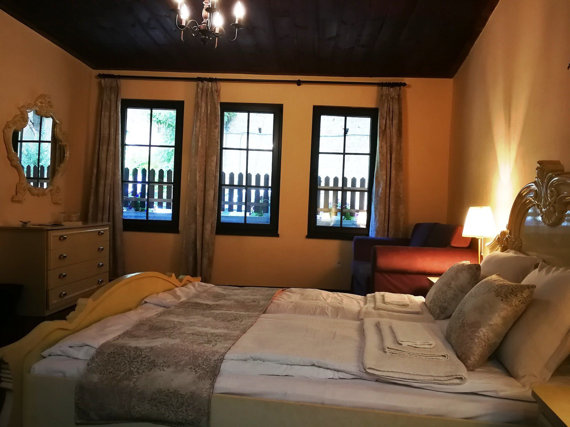 Una cama bien hecha con un edredón estampado, un aparador con cajones, dos ventanas con cortinas y una araña.