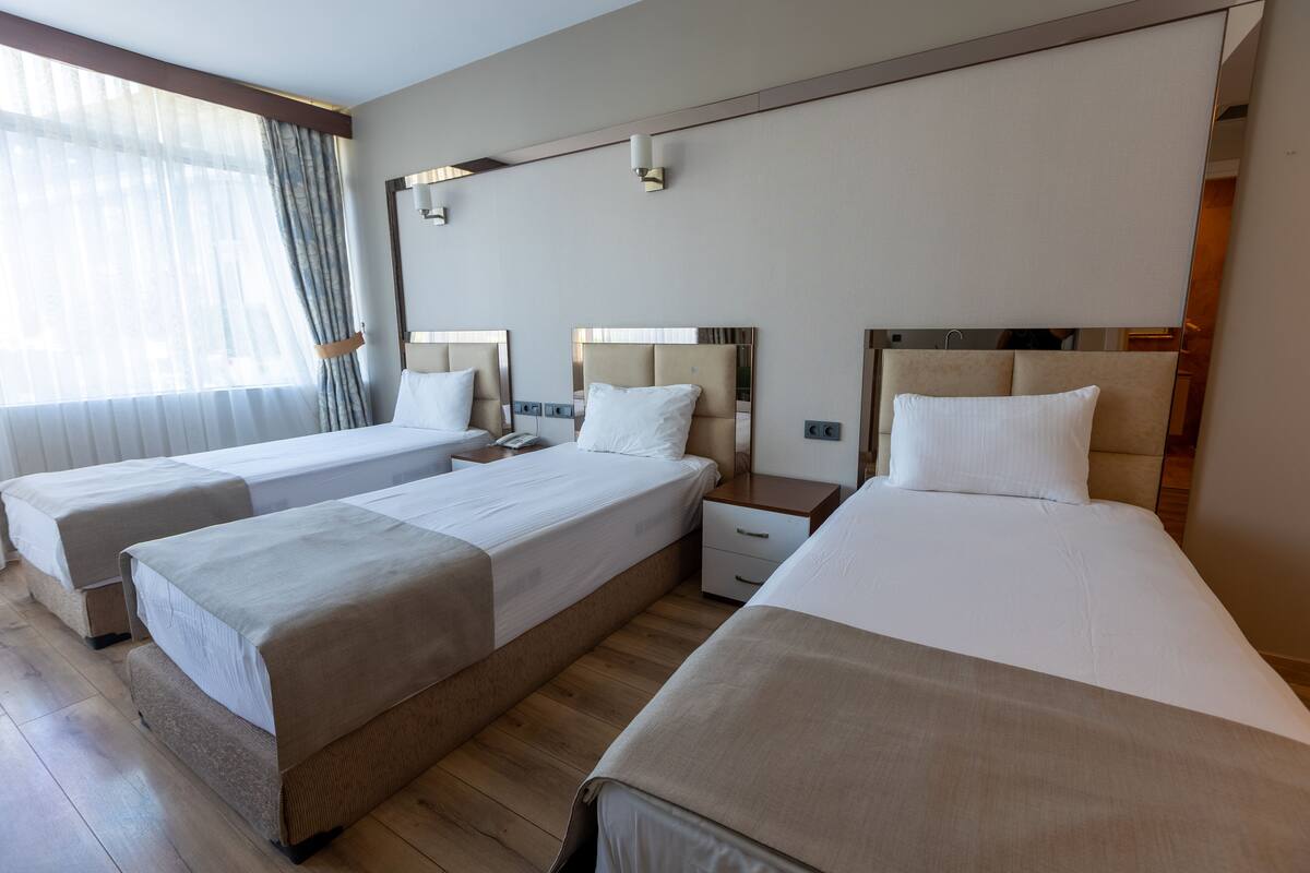 A hotel room with three single beds, wooden flooring, and a large window with sheer curtains.