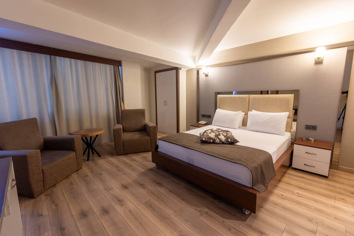 A modern hotel room with a large bed, two armchairs, a small round table, and a nightstand with a lamp.