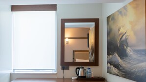 A compact room with a wooden desk, a small cabinet, a trash bin, a cushioned stool, a window with blinds, a wall-mounted mirror, and a large abstract painting. - Asia Business Suites (Istanbul)