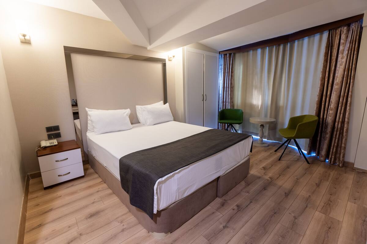 A modern hotel room with a large bed, a bedside table, a wardrobe, a small table with a chair, and a view of the outdoors through curtains.