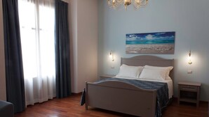 Een slaapkamer met een bed, twee nachtkastjes, een kroonluchter, een schilderij van een strand en een raam met gordijnen.