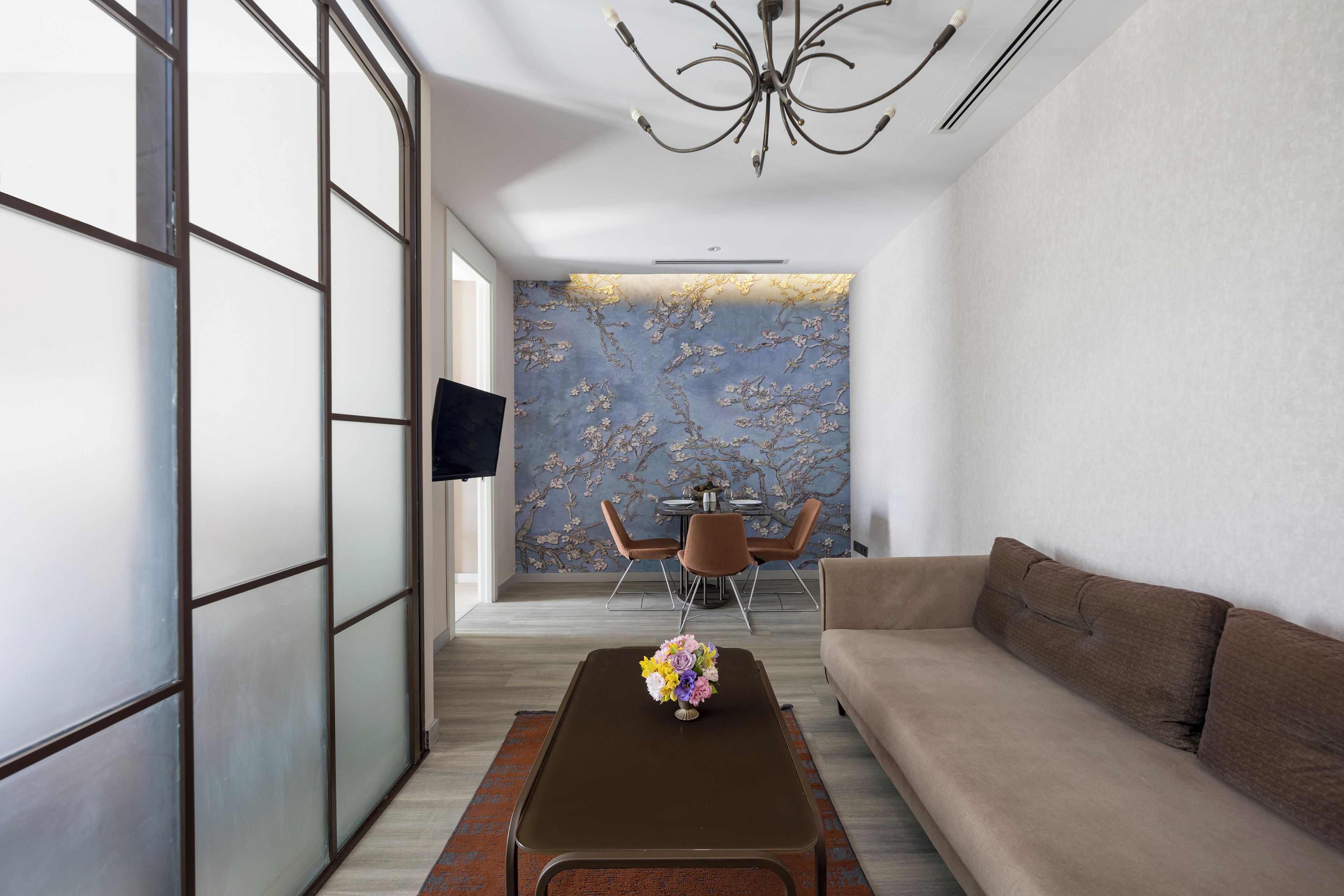 Una sala de estar moderna con un sofá, una mesa de centro con un arreglo floral, una zona de comedor con sillas y una mesa, y un televisor montado en la pared.