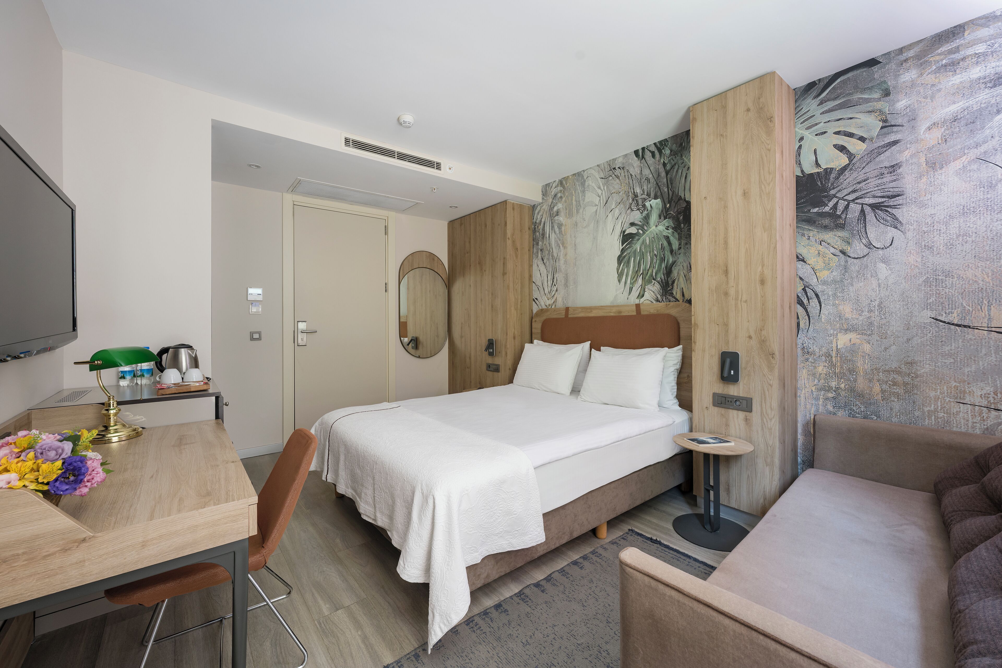 A hotel room with a bed, a desk with a lamp, a chair, a sofa, and a painting on the wall.