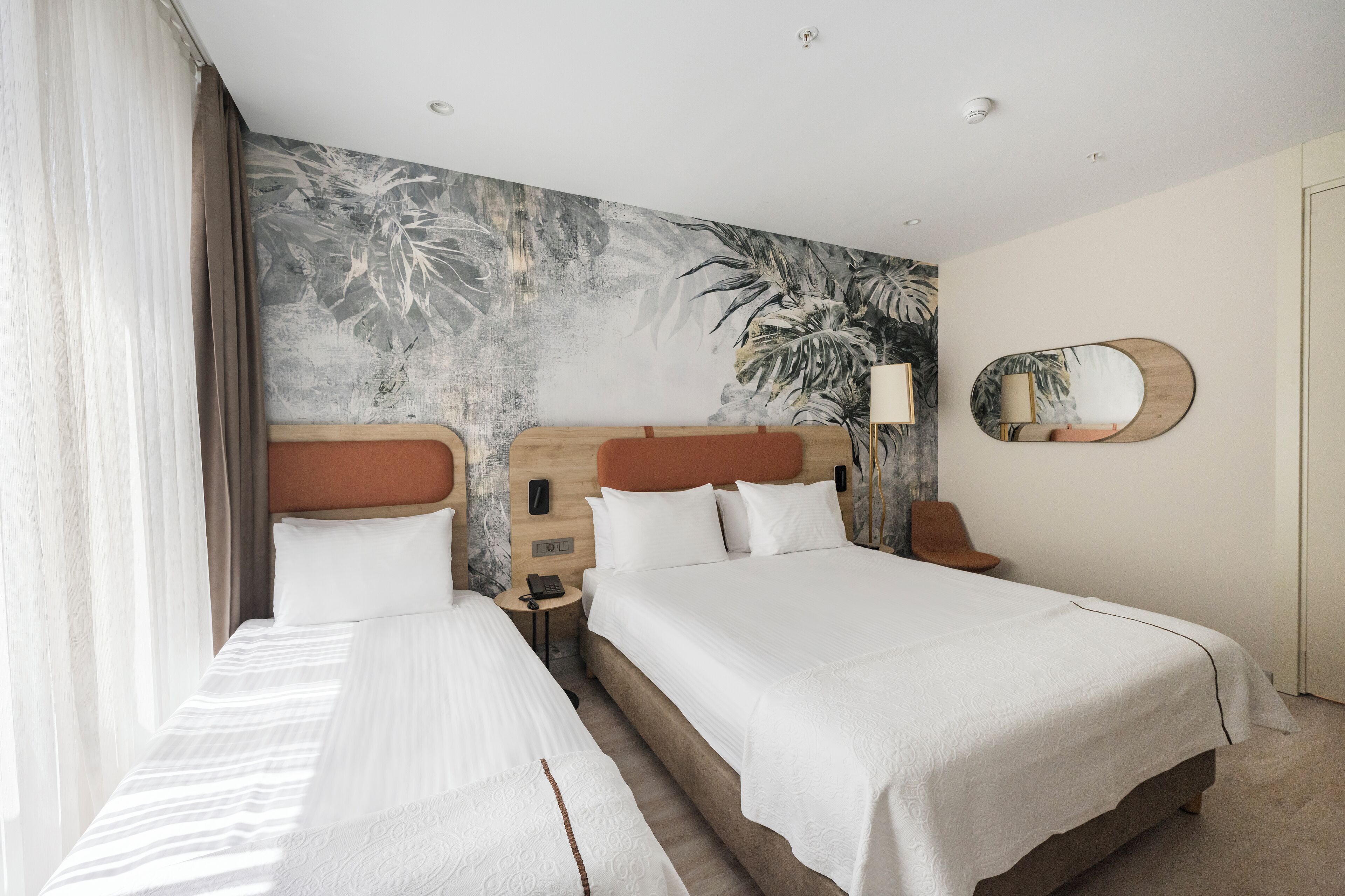 Une chambre d’hôtel avec deux lits, une grande fresque murale à motifs floraux, un miroir rond et une tête de lit en bois.