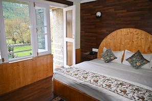 A bedroom with a wooden headboard, a bed with patterned bedding, a nightstand, and a window with a view of greenery. - Magic Himalayan Cottage (Pahalgam)