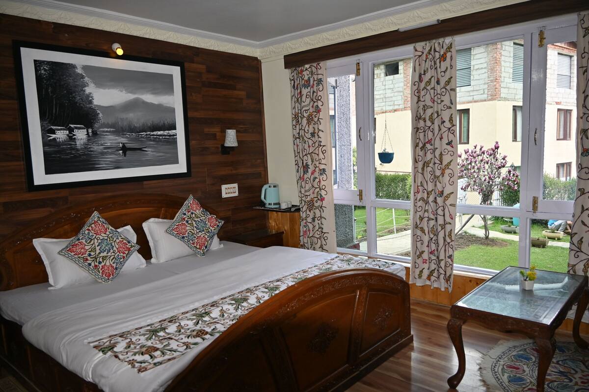Habitación de hotel con cabecera de madera, cama con almohadas estampadas, mesita de noche con lámpara y un ventanal con cortinas.