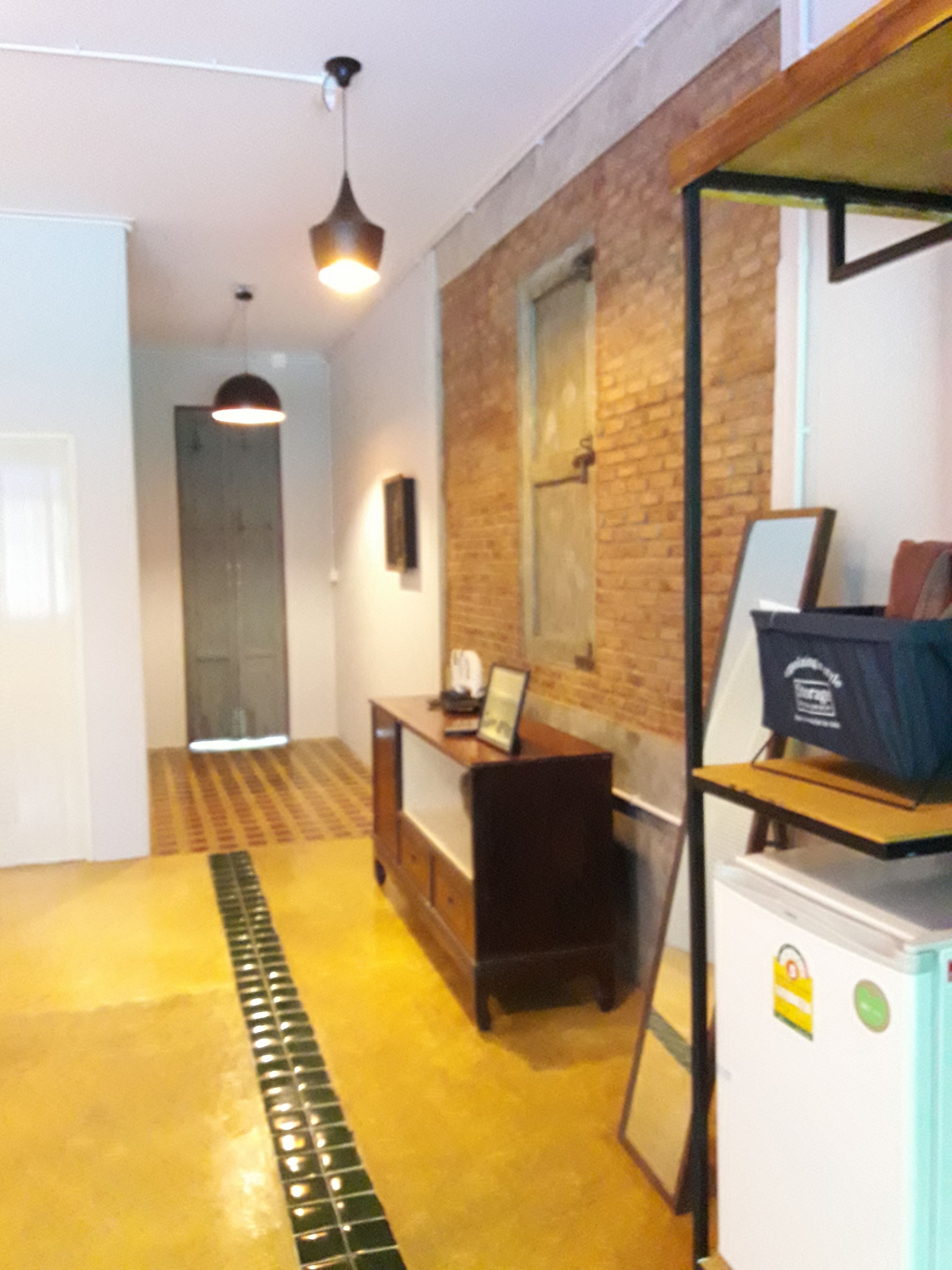 A hallway with a tiled floor, a desk with a computer, a refrigerator, and a hanging light fixture.