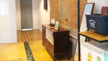 A hallway with a tiled floor, a desk with a computer, a refrigerator, and a hanging light fixture.