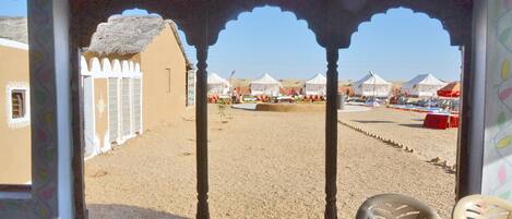 A view from a window with an arched doorway, leading to a sandy area with tents and a clear sky.