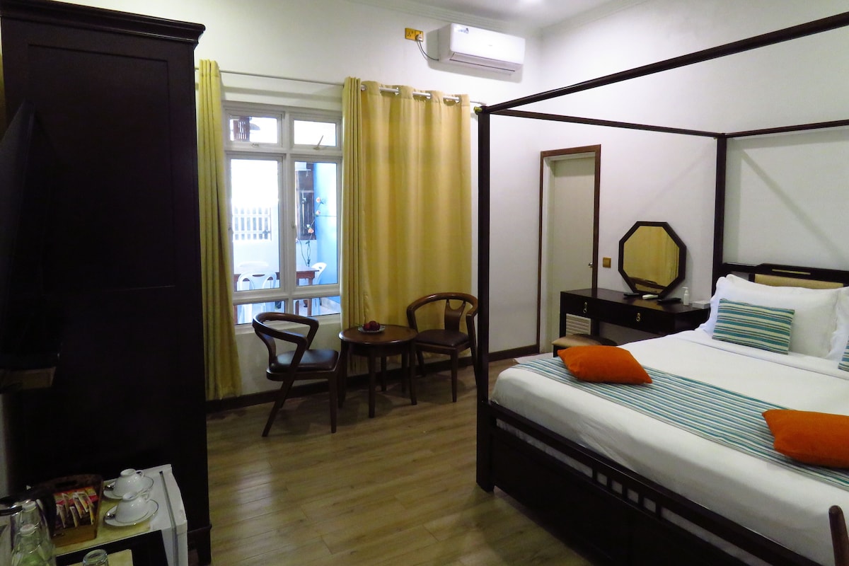 A four-poster bed with a wooden frame, a small bedside table with a mirror, and two chairs in a room with wooden flooring and a window with curtains.