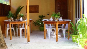 Um pátio externo com mesas de madeira e cadeiras de plástico branco, rodeado por vasos de plantas.