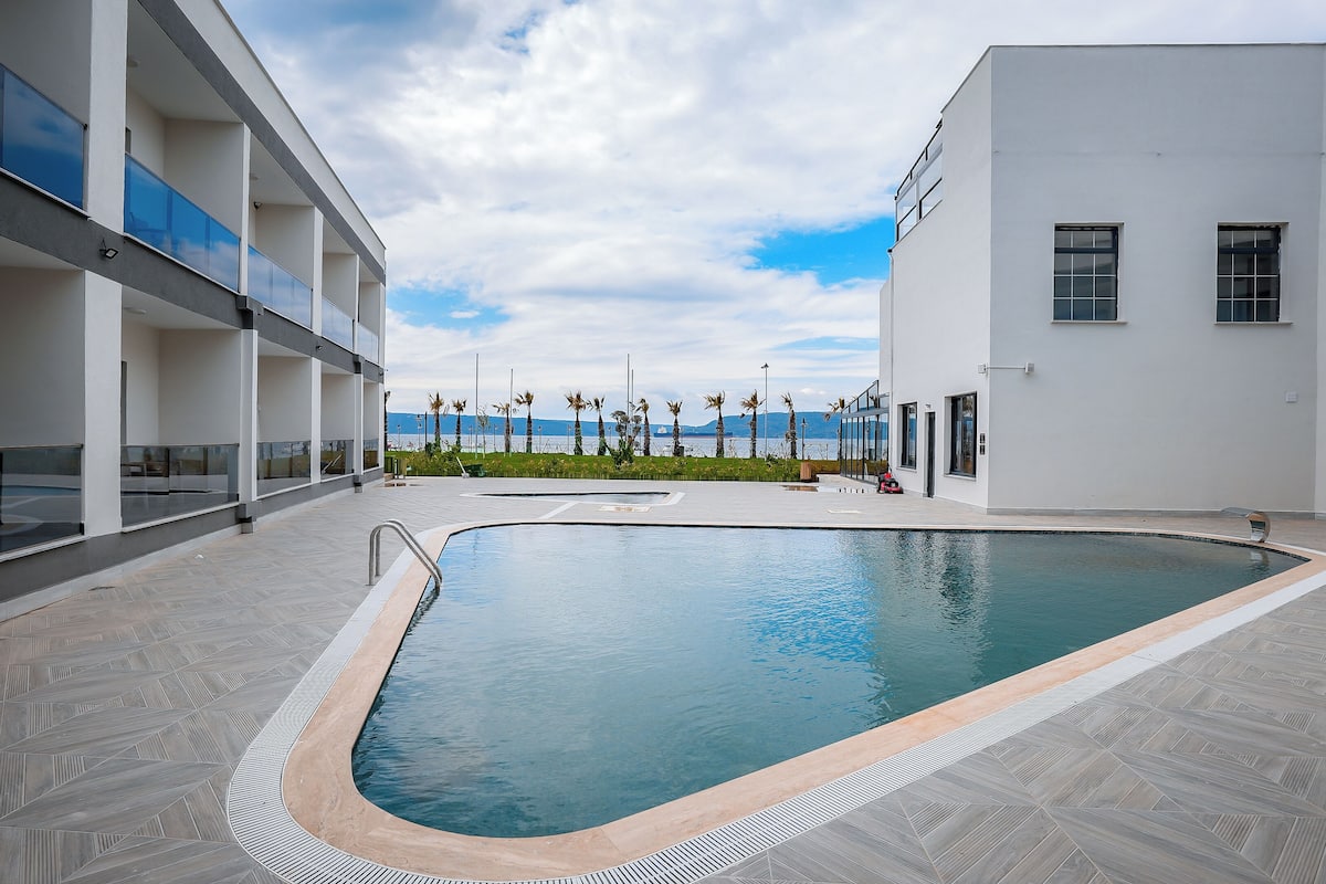 A modern pool area with a clear view of the sea, a building with balconies, and a tiled floor.