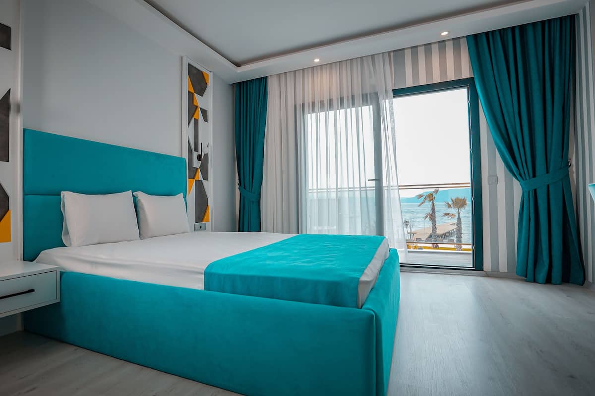 A modern hotel room with a large bed, a nightstand, and a view of the sea through a balcony.