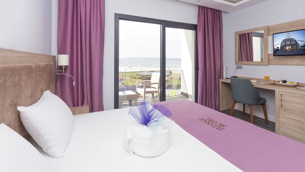 A hotel room with a bed, a desk, a chair, a TV, and a view of the beach. - Sunsan Hotel (Çanakkale)