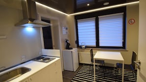 A compact kitchen with a stove, sink, and a small refrigerator. There is a window with blinds and a radiator below it. - Ferienzimmer 1 Saarlouis (Saarlouis)